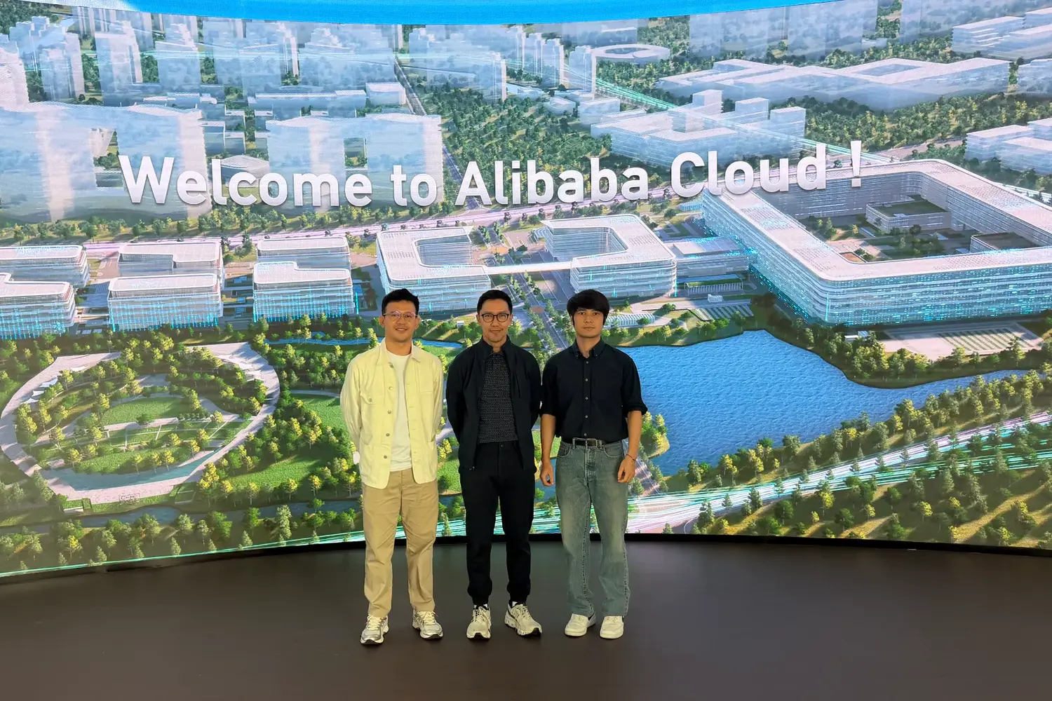 Three men standing on a dark stage in front of a large curved screen that reads “Welcome to Alibaba Cloud!” over an aerial rendering of a campus of white buildings, trees and a lake.
